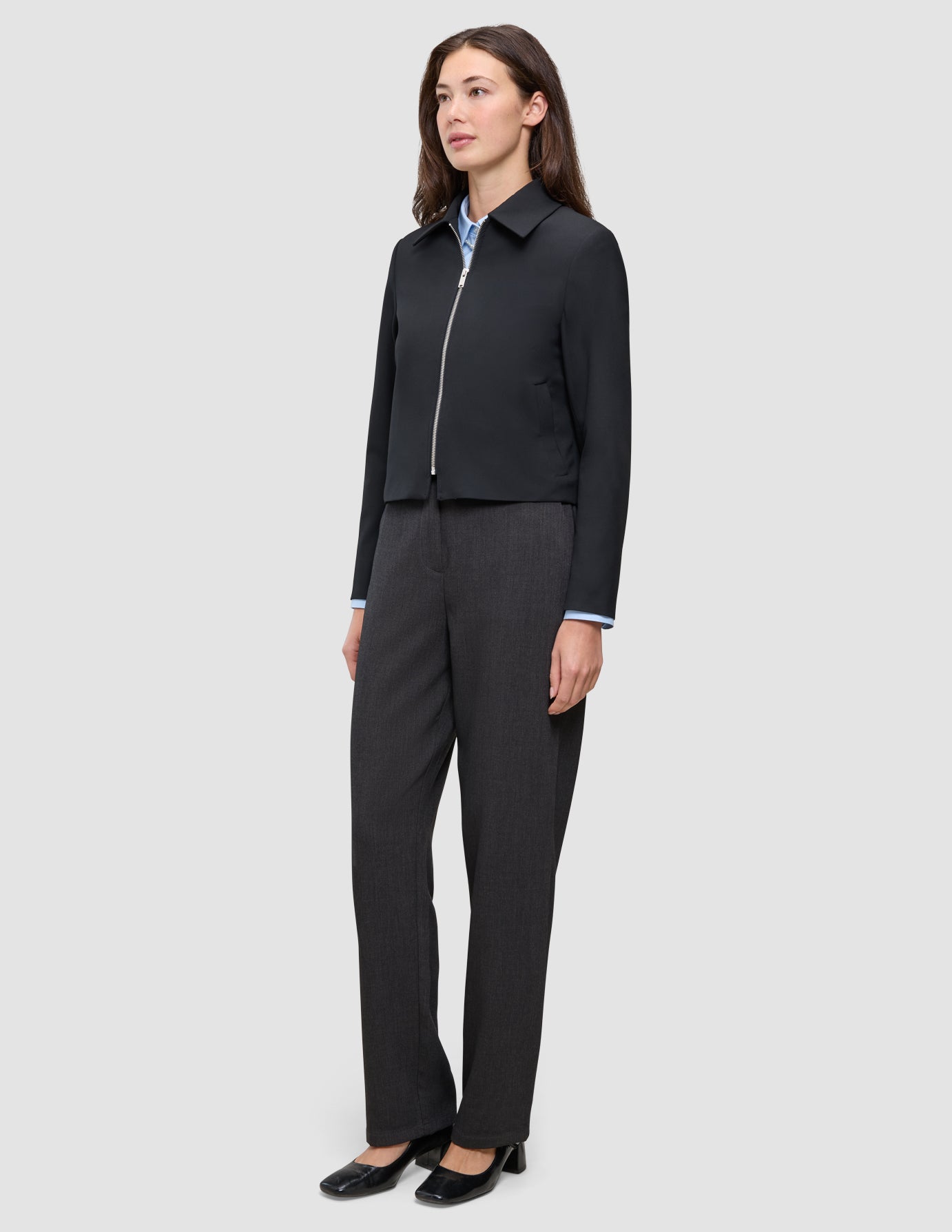 Eine Person, die eine schwarze Essential Short Zip Jacket über einem hellblauen Hemd mit Kragen und einer dunkelgrauen Hose trägt; das Design besticht durch eine schlichte, skandinavische Ästhetik.